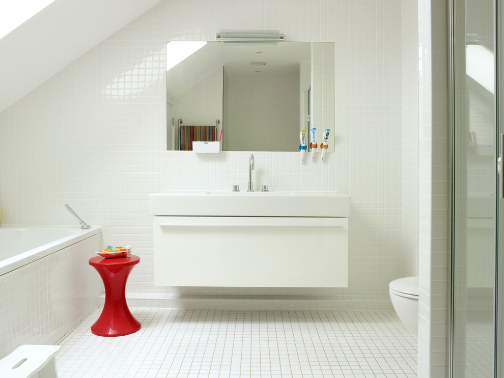 This is an example of a mid-sized contemporary kids bathroom in London with flat-panel cabinets, white cabinets, a drop-in tub, white tile, ceramic tile, white walls, mosaic tile floors, a curbless shower, a wall-mount toilet and a wall-mount sink.