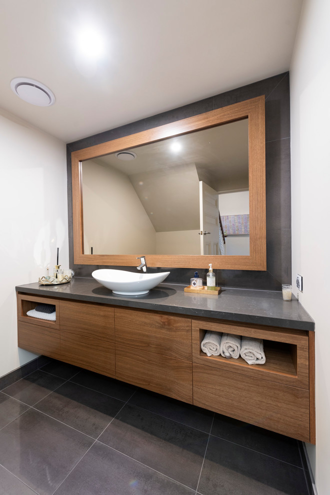 Photo of a mid-sized contemporary powder room in Melbourne with furniture-like cabinets, medium wood cabinets, a two-piece toilet, black tile, porcelain tile, white walls, porcelain floors, a vessel sink, engineered quartz benchtops, black floor and black benchtops.