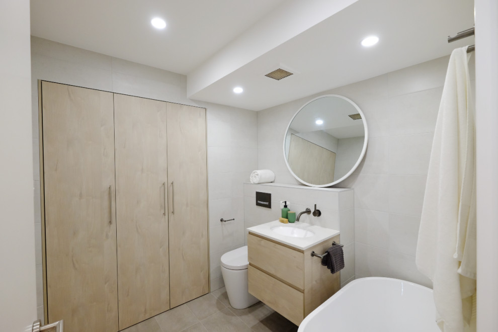 Idée de décoration pour une petite salle de bain urbaine en bois clair avec un placard à porte plane, une baignoire indépendante, une douche ouverte, WC suspendus, un carrelage blanc, des carreaux de porcelaine, un mur blanc, un sol en carrelage de porcelaine, un lavabo posé, un plan de toilette en quartz, un sol gris, aucune cabine, un plan de toilette blanc, buanderie, meuble simple vasque et meuble-lavabo suspendu.