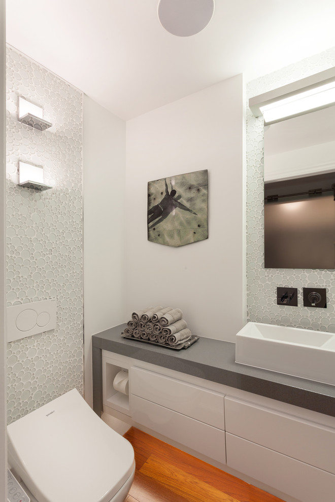 This is an example of a small contemporary powder room in Ottawa with flat-panel cabinets, white cabinets, a wall-mount toilet, ceramic tile, white walls, medium hardwood floors, a vessel sink and engineered quartz benchtops.