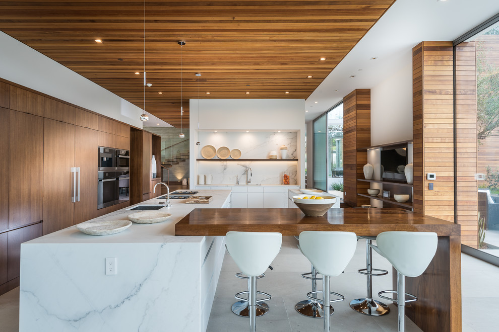 Contemporary l-shaped kitchen in Los Angeles with an undermount sink, flat-panel cabinets, dark wood cabinets, stainless steel appliances, multiple islands, white floor and white benchtop.