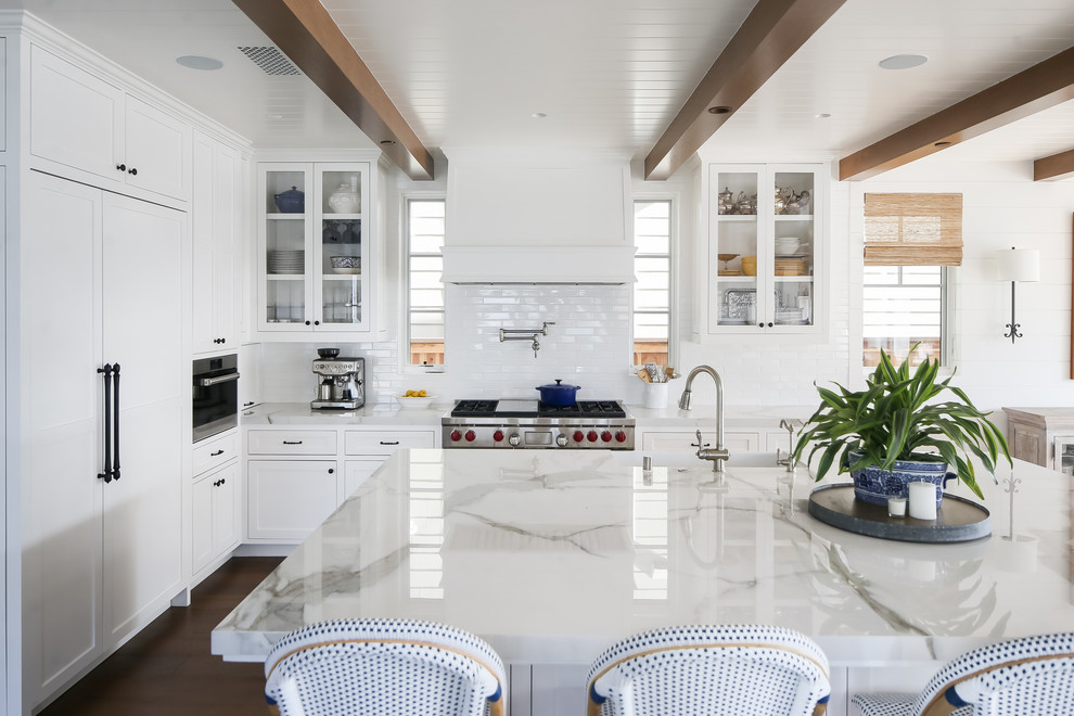 This is an example of a beach style l-shaped kitchen in Orange County with glass-front cabinets, white cabinets, white splashback, subway tile splashback, stainless steel appliances, with island, brown floor, white benchtop and dark hardwood floors.