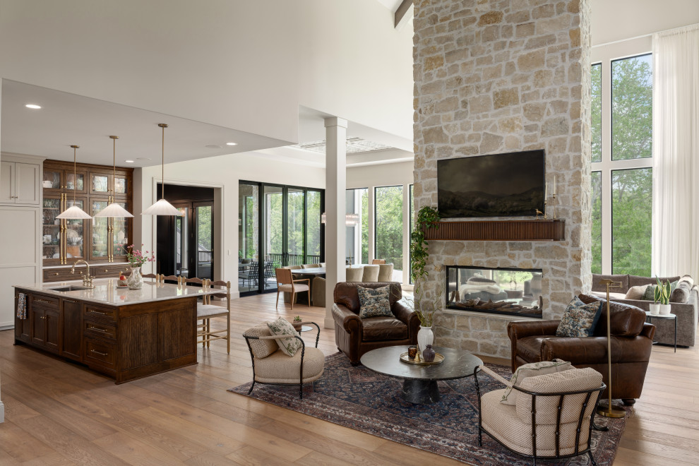 French country open concept medium tone wood floor, brown floor and vaulted ceiling living room photo in Kansas City with white walls, a two-sided fireplace, a stone fireplace and a wall-mounted tv