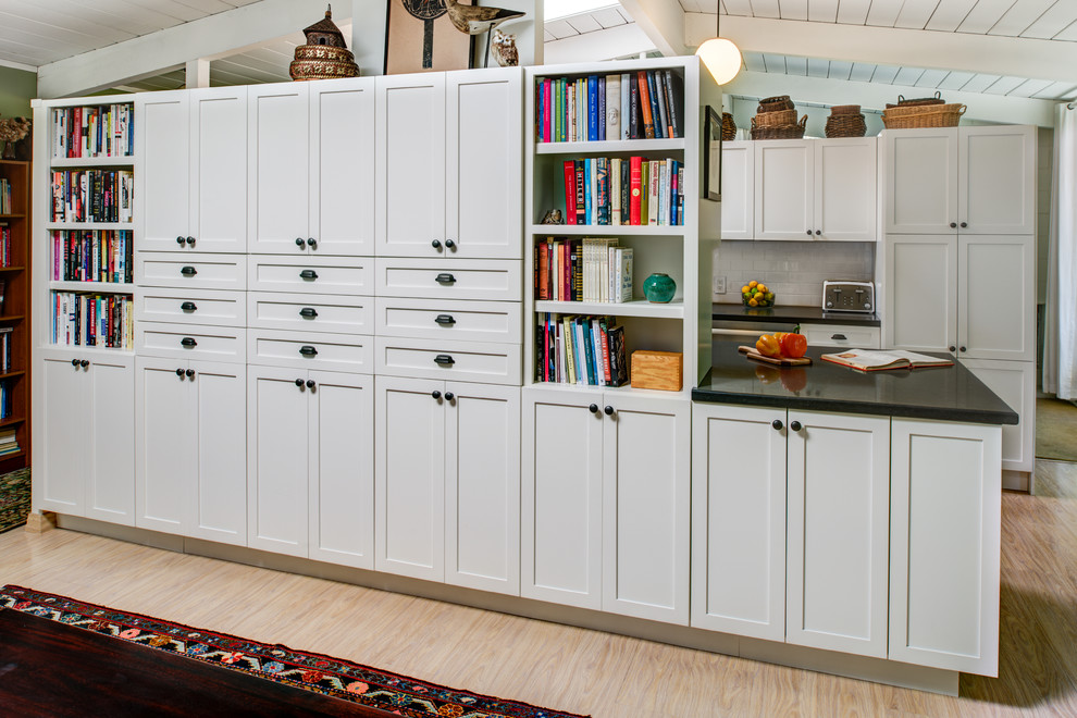 This is an example of a small arts and crafts galley kitchen pantry in San Francisco with a farmhouse sink, shaker cabinets, white cabinets, solid surface benchtops, white splashback, porcelain splashback, stainless steel appliances, light hardwood floors and with island.
