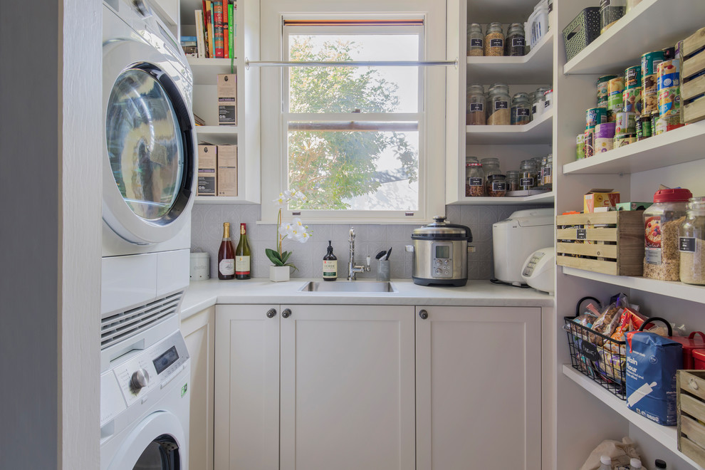 Inspiration for a mid-sized transitional u-shaped utility room in Sydney with shaker cabinets, white cabinets, quartz benchtops, grey splashback, ceramic splashback, white benchtop, a drop-in sink and a stacked washer and dryer.