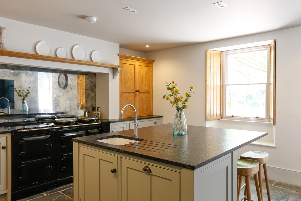 Country l-shaped kitchen in Cornwall with an undermount sink, shaker cabinets, green cabinets, mirror splashback, black appliances, with island and black benchtop.