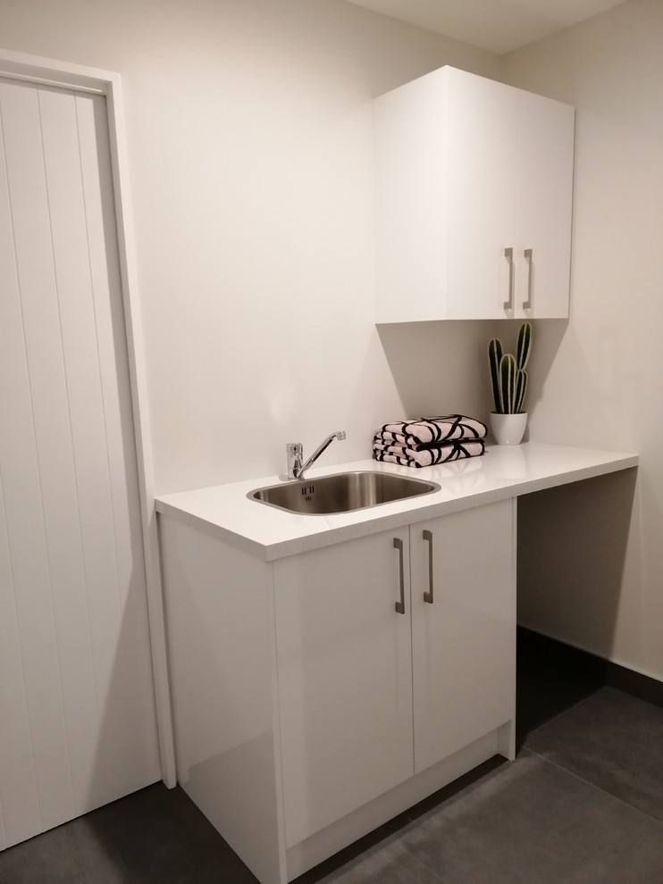 Small contemporary single-wall dedicated laundry room in Christchurch with a drop-in sink, flat-panel cabinets, white cabinets, laminate benchtops, white walls, linoleum floors, an integrated washer and dryer, grey floor and white benchtop.