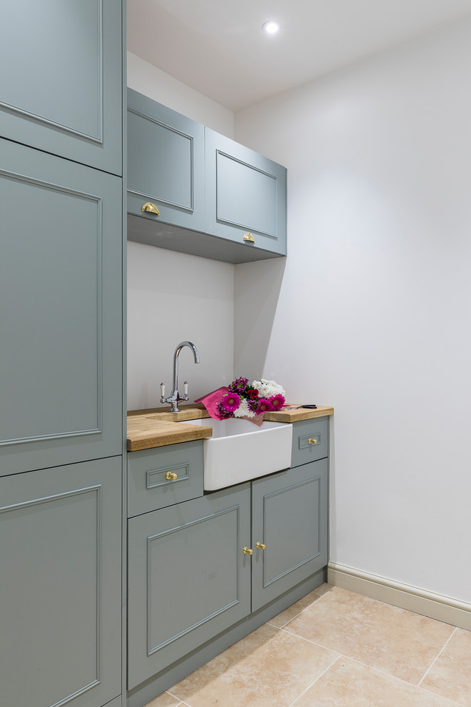 Photo of a small traditional single-wall dedicated laundry room in London with a farmhouse sink, shaker cabinets, green cabinets, wood benchtops, grey walls, slate floors, beige floor and brown benchtop.