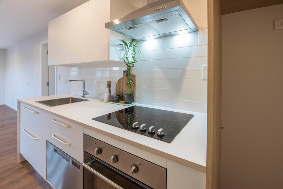 This is an example of a small scandinavian single-wall separate kitchen in Christchurch with a single-bowl sink, flat-panel cabinets, white cabinets, laminate benchtops, white splashback, subway tile splashback, stainless steel appliances, vinyl floors, no island, brown floor and white benchtop.
