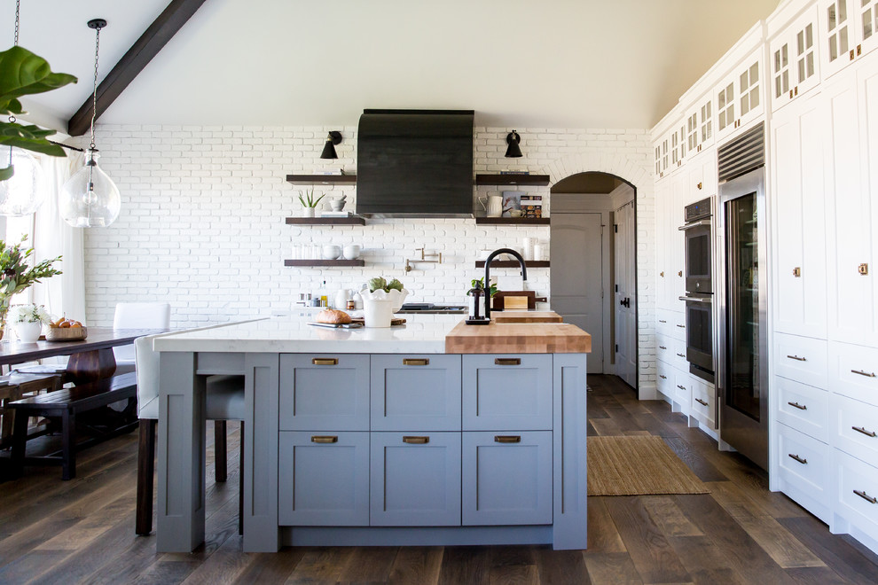 Design ideas for a mid-sized country l-shaped eat-in kitchen in Salt Lake City with a farmhouse sink, shaker cabinets, white cabinets, quartz benchtops, white splashback, brick splashback, black appliances, medium hardwood floors and with island.