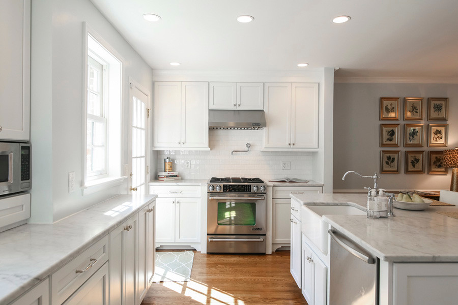 Photo of a mid-sized traditional galley kitchen in DC Metro with a farmhouse sink, recessed-panel cabinets, white cabinets, marble benchtops, white splashback, ceramic splashback, stainless steel appliances, light hardwood floors and a peninsula.