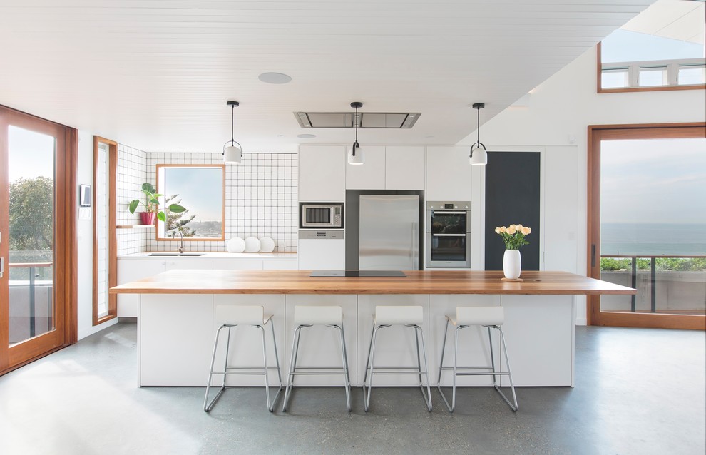 Inspiration for a beach style galley open plan kitchen in Newcastle - Maitland with an undermount sink, flat-panel cabinets, white cabinets, wood benchtops, white splashback, stainless steel appliances, concrete floors, with island, grey floor and brown benchtop.