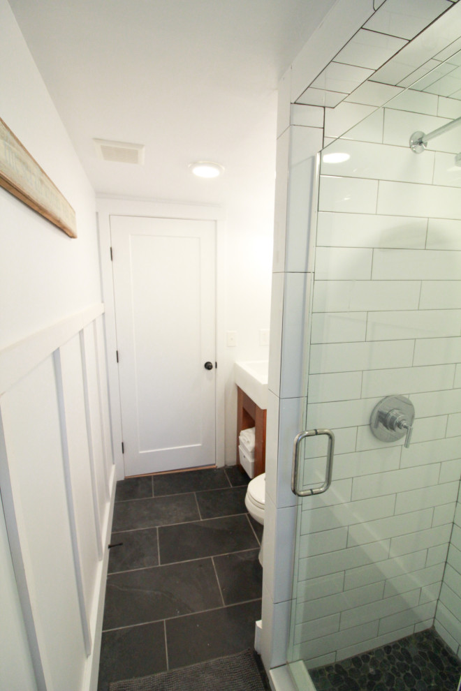 Inredning av ett maritimt litet vit vitt en-suite badrum, med öppna hyllor, bruna skåp, en dusch i en alkov, en toalettstol med hel cisternkåpa, vit kakel, keramikplattor, vita väggar, skiffergolv, ett avlångt handfat, bänkskiva i akrylsten, svart golv och dusch med gångjärnsdörr
