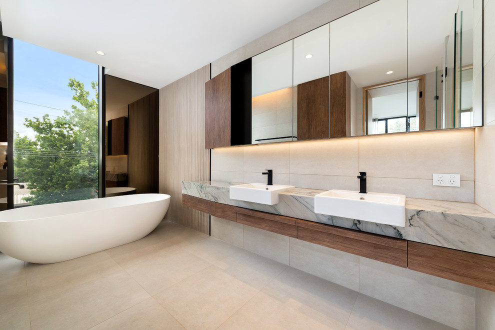 Modern master bathroom in Melbourne with flat-panel cabinets, medium wood cabinets, a freestanding tub, beige tile, a vessel sink, beige floor and grey benchtops.