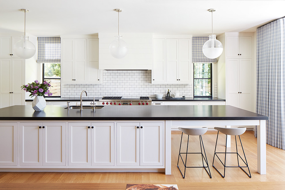 Example of a large transitional galley light wood floor and beige floor open concept kitchen design in San Francisco with an undermount sink, white cabinets, white backsplash, subway tile backsplash, stainless steel appliances, an island, shaker cabinets, solid surface countertops and black countertops