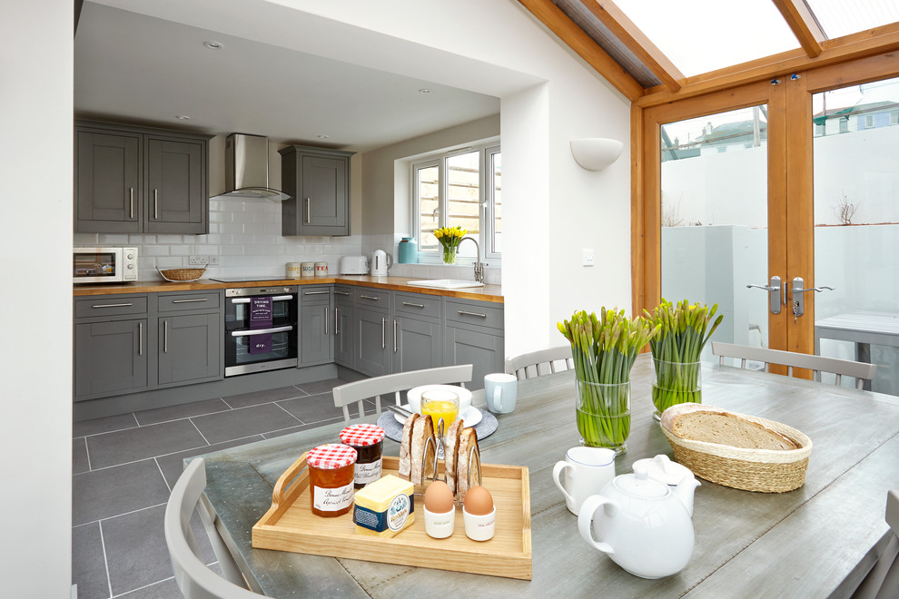 Photo of a beach style eat-in kitchen in London with a drop-in sink, shaker cabinets, grey cabinets, wood benchtops, white splashback, subway tile splashback, stainless steel appliances, no island and grey floor.