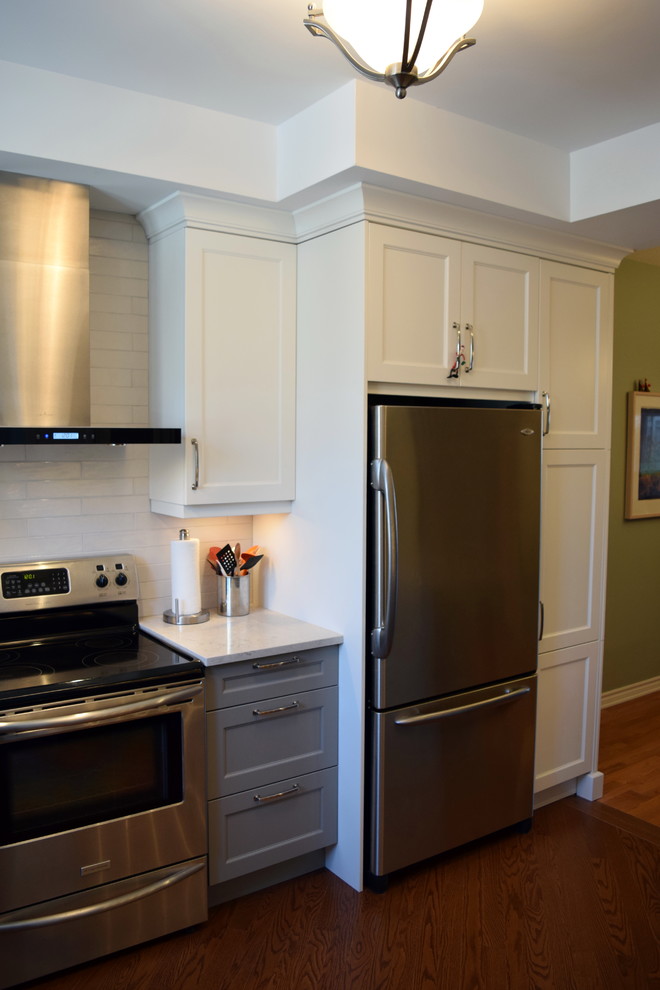 Example of a mid-sized transitional l-shaped medium tone wood floor eat-in kitchen design in Toronto with an undermount sink, shaker cabinets, white cabinets, quartz countertops, white backsplash, subway tile backsplash, stainless steel appliances and a peninsula