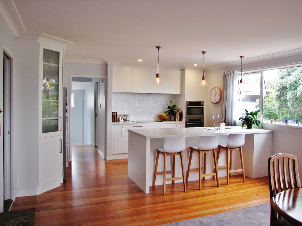 Large single-wall eat-in kitchen in Auckland with a triple-bowl sink, raised-panel cabinets, white cabinets, granite benchtops, white splashback, metal splashback, stainless steel appliances, medium hardwood floors, with island, brown floor and white benchtop.