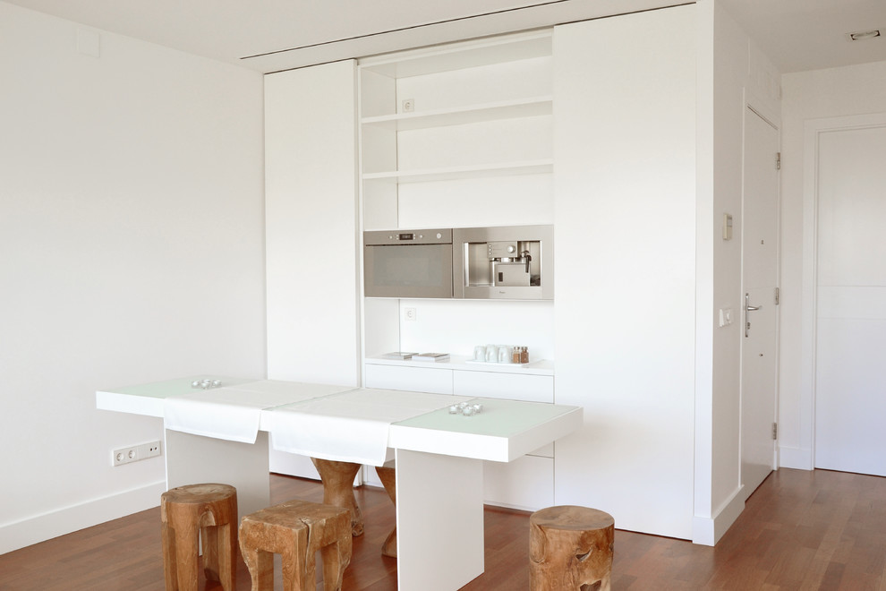 This is an example of a small scandinavian single-wall open plan kitchen in Barcelona with flat-panel cabinets, white cabinets, stainless steel appliances, with island, solid surface benchtops, white splashback and medium hardwood floors.