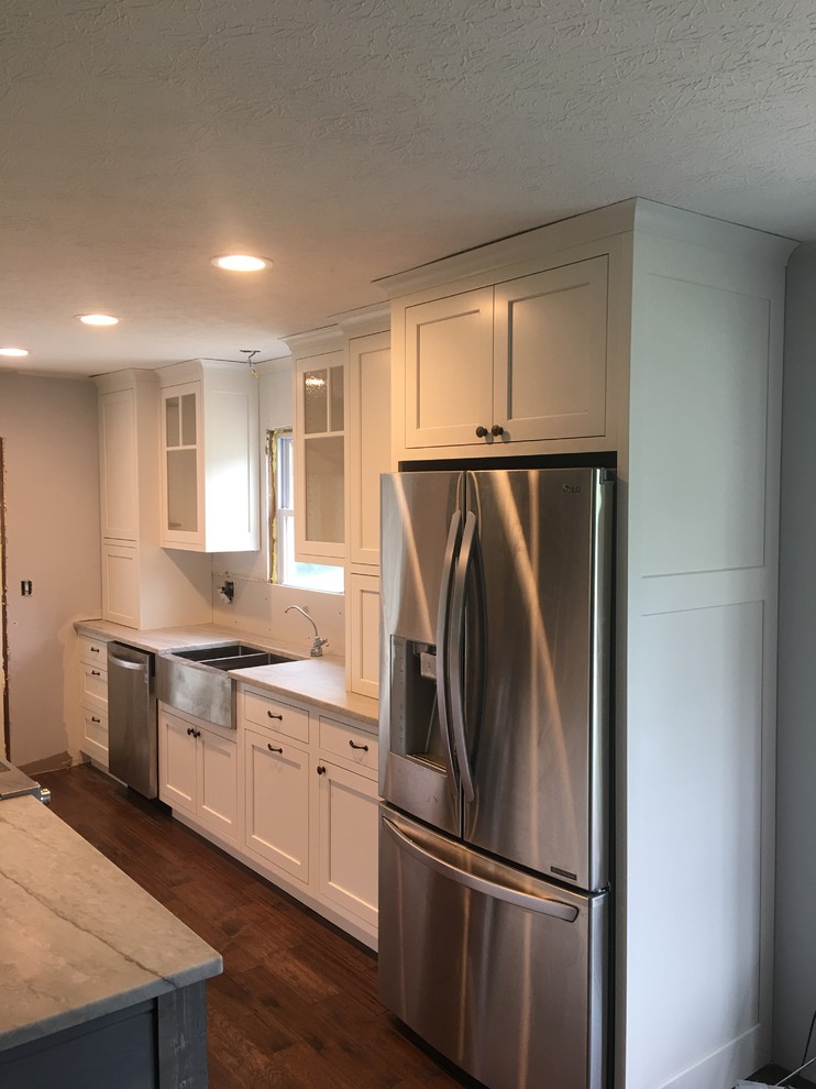 This is an example of a mid-sized arts and crafts single-wall kitchen in Columbus with a farmhouse sink, shaker cabinets, white cabinets, quartzite benchtops, stainless steel appliances, with island and brown floor.