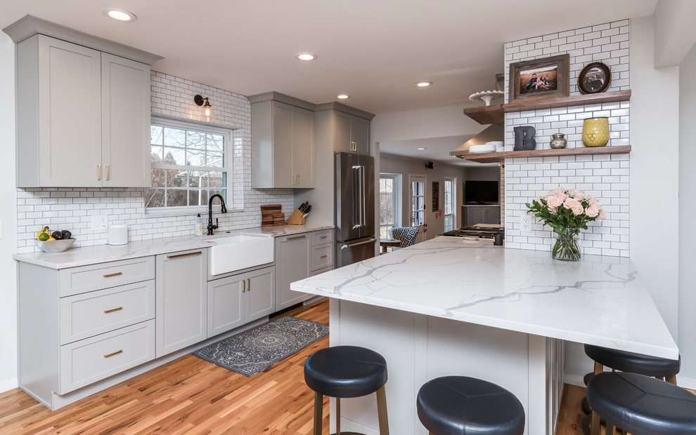 Eat-in kitchen - mid-sized transitional galley medium tone wood floor eat-in kitchen idea in Denver with a farmhouse sink, shaker cabinets, gray cabinets, quartz countertops, white backsplash, porcelain backsplash, stainless steel appliances, a peninsula and white countertops