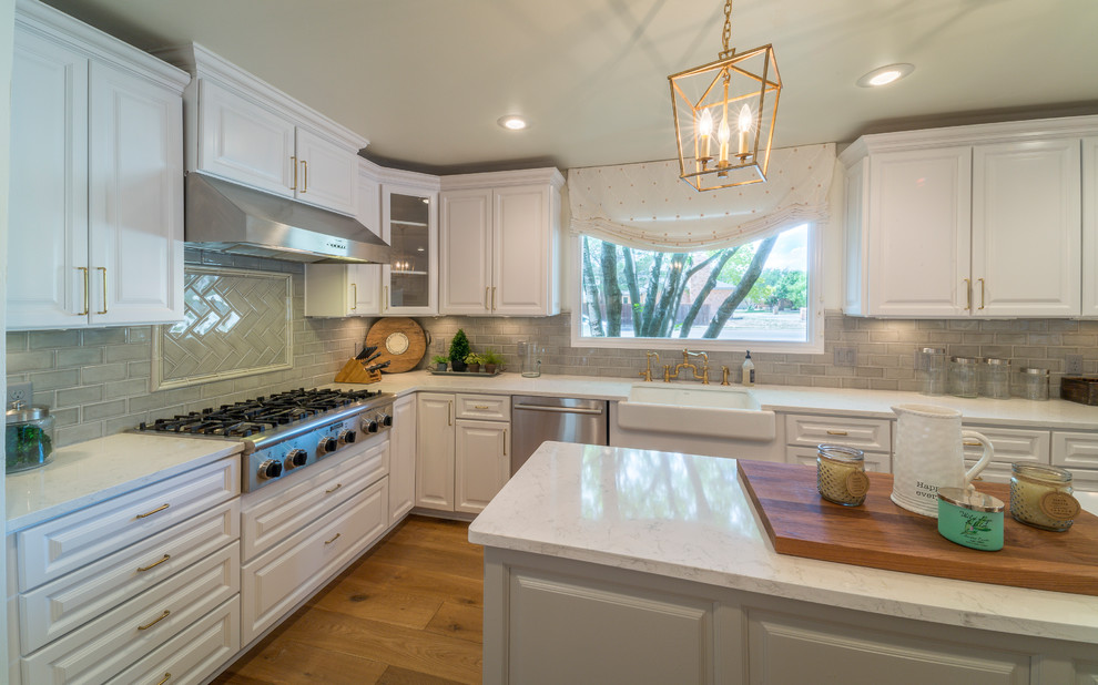 Example of a large transitional u-shaped medium tone wood floor and brown floor enclosed kitchen design with a farmhouse sink, raised-panel cabinets, white cabinets, quartz countertops, gray backsplash, glass tile backsplash, stainless steel appliances and an island