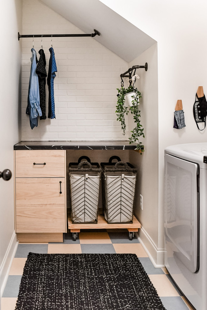 Inspiration for a mid-sized industrial l-shaped dedicated laundry room in Atlanta with flat-panel cabinets, light wood cabinets, laminate benchtops, white walls, linoleum floors, a side-by-side washer and dryer, multi-coloured floor, black benchtop and brick walls.