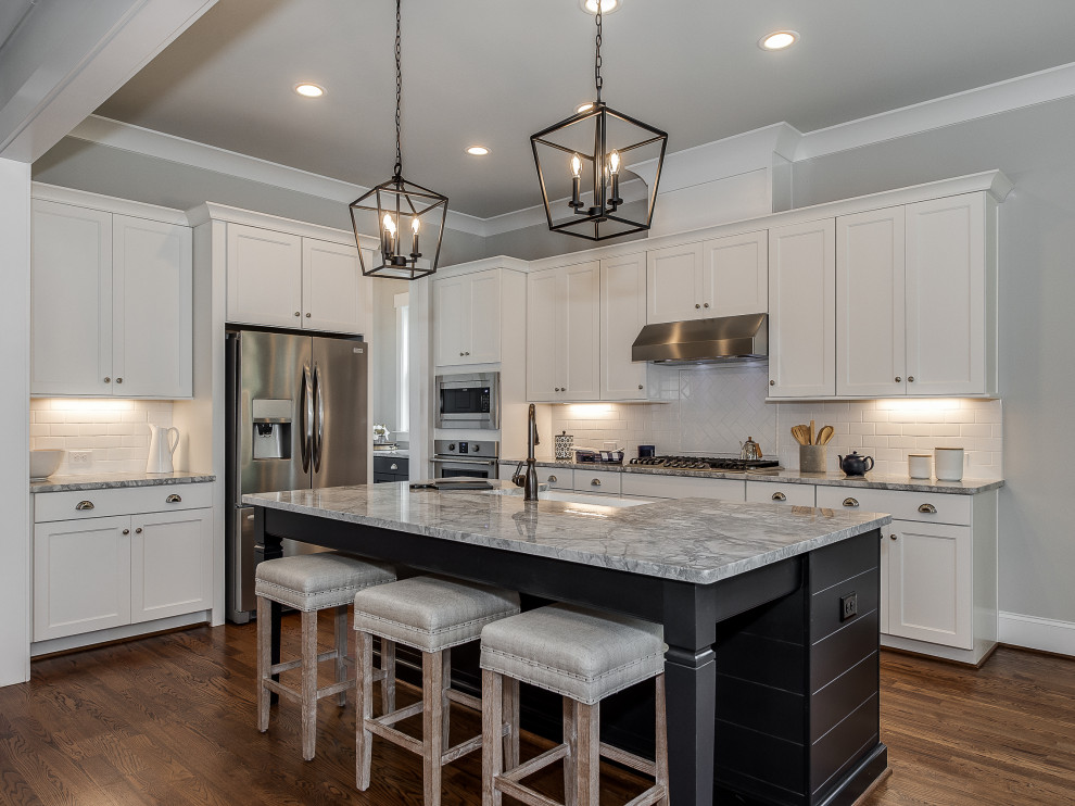 Expansive transitional l-shaped eat-in kitchen in Charlotte with a farmhouse sink, shaker cabinets, white cabinets, marble benchtops, white splashback, subway tile splashback, stainless steel appliances, medium hardwood floors, with island, brown floor and grey benchtop.