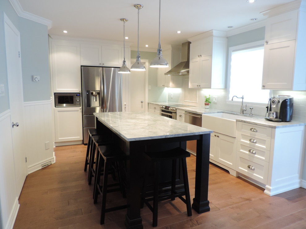 Photo of a mid-sized transitional l-shaped eat-in kitchen in Montreal with a farmhouse sink, shaker cabinets, white cabinets, granite benchtops, white splashback, ceramic splashback, stainless steel appliances, light hardwood floors and with island.