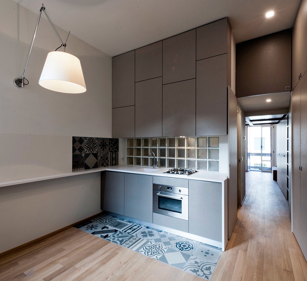 This is an example of a small modern single-wall eat-in kitchen in Milan with an integrated sink, flat-panel cabinets, grey cabinets, solid surface benchtops, white splashback, glass sheet splashback, stainless steel appliances and ceramic floors.