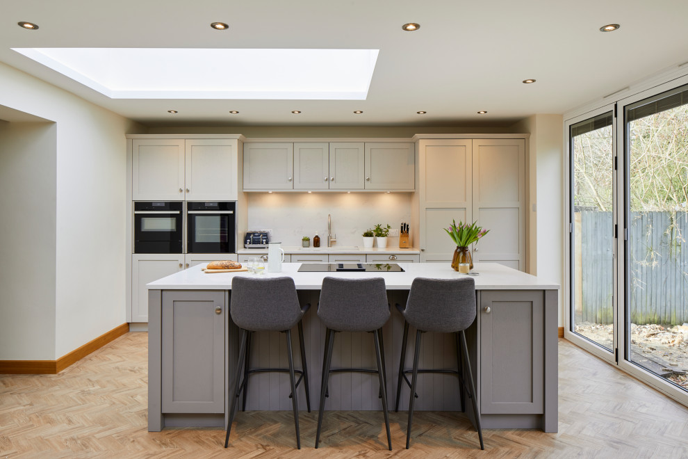 Mid-sized transitional single-wall eat-in kitchen in London with an undermount sink, shaker cabinets, grey cabinets, quartzite benchtops, white splashback, engineered quartz splashback, panelled appliances, medium hardwood floors, with island, brown floor and white benchtop.