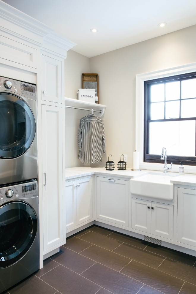 This is an example of a large traditional u-shaped dedicated laundry room in Toronto with a farmhouse sink, recessed-panel cabinets, white cabinets, marble benchtops, beige walls, brick floors, a stacked washer and dryer, black floor and white benchtop.