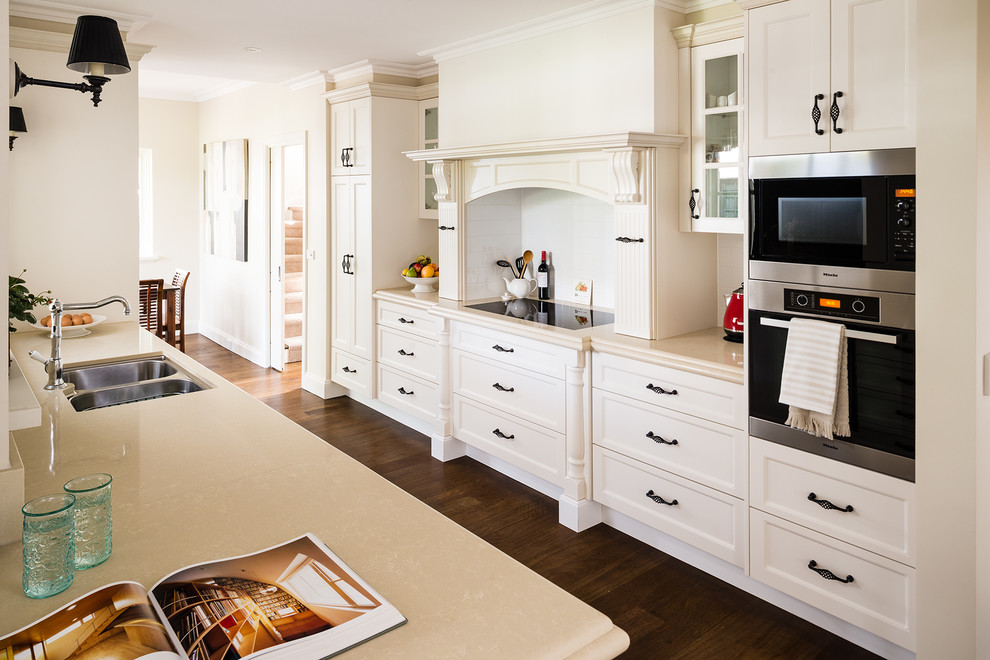 This is an example of a large modern galley eat-in kitchen in Melbourne with a double-bowl sink, beaded inset cabinets, white cabinets, quartz benchtops, white splashback, ceramic splashback, black appliances, medium hardwood floors and a peninsula.