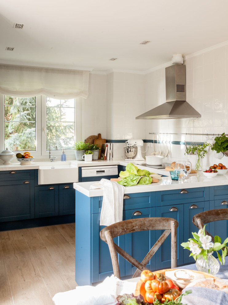 Design ideas for a transitional u-shaped eat-in kitchen in Bilbao with a farmhouse sink, shaker cabinets, blue cabinets, white splashback, light hardwood floors, a peninsula, brown floor and white benchtop.