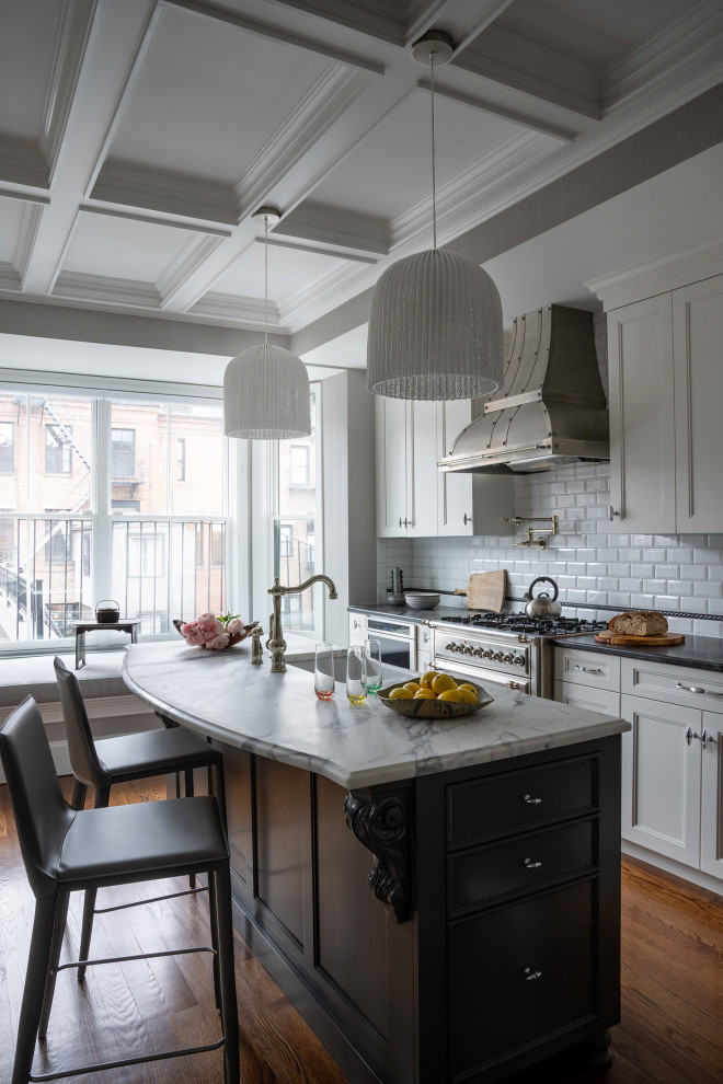 Mid-sized elegant galley medium tone wood floor, brown floor and coffered ceiling kitchen photo in Boston with a farmhouse sink, white cabinets, white backsplash, stainless steel appliances, an island, recessed-panel cabinets, subway tile backsplash and black countertops