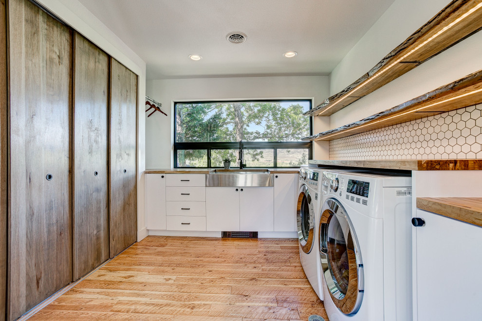 Design ideas for a country l-shaped dedicated laundry room in Denver with a farmhouse sink, flat-panel cabinets, white cabinets, wood benchtops, white walls, medium hardwood floors, a stacked washer and dryer, brown floor and brown benchtop.