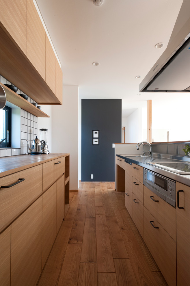 Example of a danish single-wall medium tone wood floor and brown floor open concept kitchen design with a single-bowl sink, flat-panel cabinets, medium tone wood cabinets, stainless steel countertops, white backsplash, a peninsula and brown countertops
