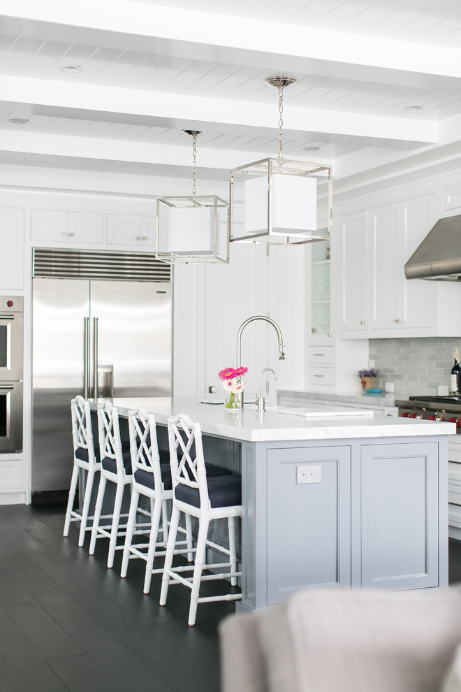 This is an example of a large beach style l-shaped open plan kitchen in Orange County with a farmhouse sink, white cabinets, marble benchtops, grey splashback, ceramic splashback, stainless steel appliances, dark hardwood floors, with island and shaker cabinets.