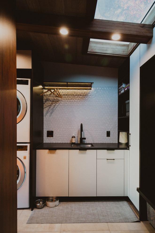 This is an example of a small modern single-wall dedicated laundry room in San Francisco with a single-bowl sink, flat-panel cabinets, white cabinets, white splashback, mosaic tile splashback, white walls, a stacked washer and dryer, beige floor, black benchtop and wood.