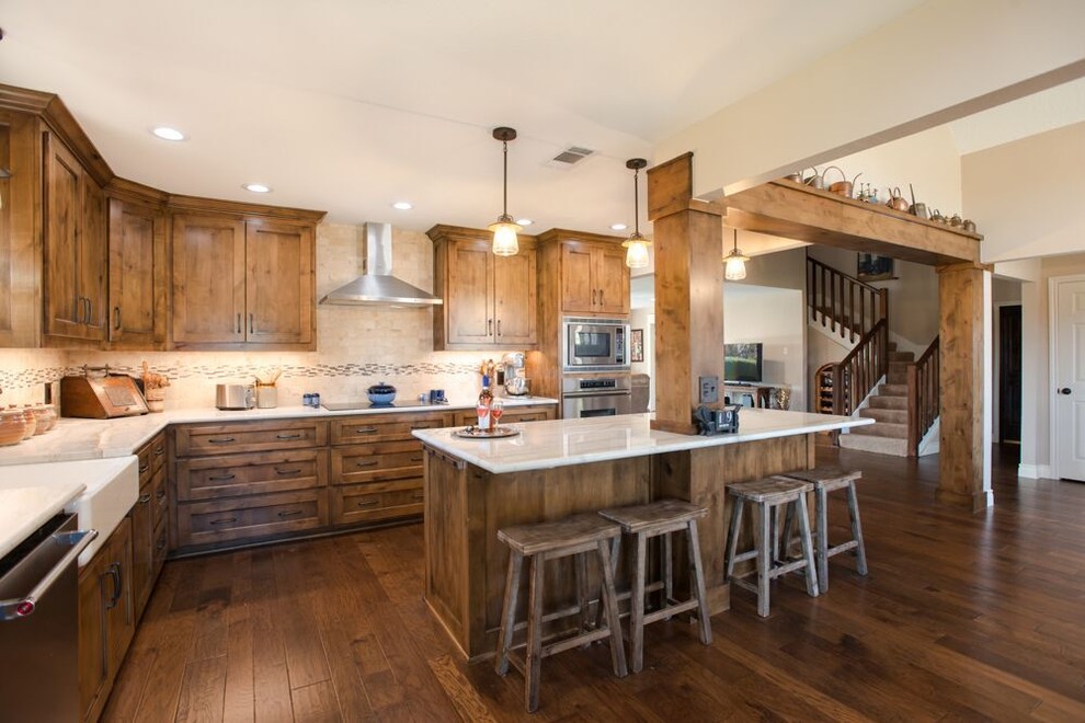 Large transitional l-shaped brown floor and medium tone wood floor eat-in kitchen photo in Dallas with a farmhouse sink, recessed-panel cabinets, medium tone wood cabinets, multicolored backsplash, an island, quartzite countertops, travertine backsplash, stainless steel appliances and white countertops
