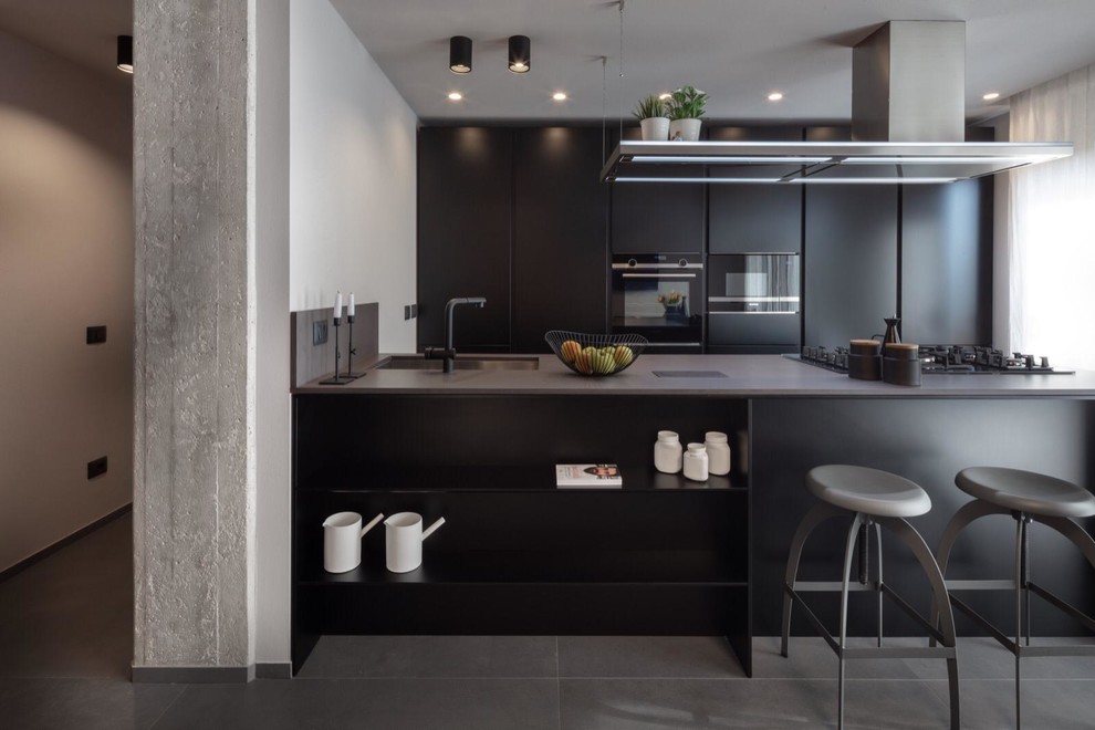 Photo of a mid-sized industrial galley kitchen in Other with an undermount sink, flat-panel cabinets, black cabinets, quartz benchtops, black appliances, porcelain floors, grey floor, grey benchtop and a peninsula.