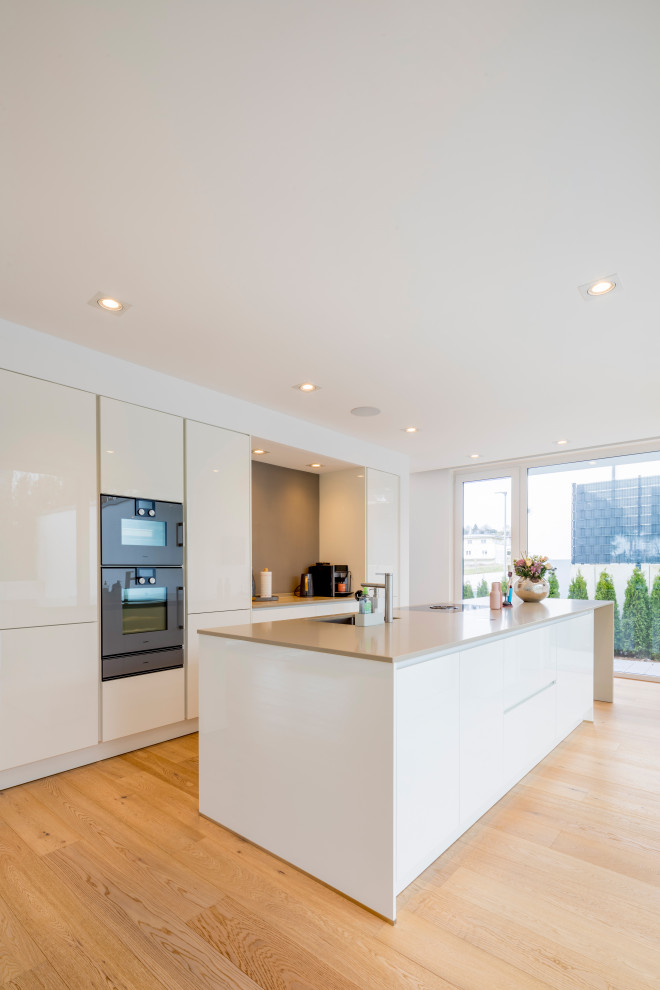 Large contemporary single-wall open plan kitchen in Dusseldorf with a drop-in sink, flat-panel cabinets, white cabinets, quartzite benchtops, grey splashback, black appliances, medium hardwood floors, with island, brown floor and beige benchtop.