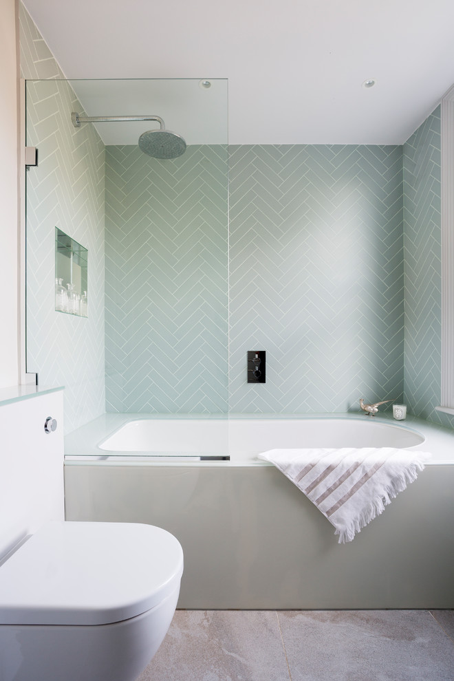This is an example of a contemporary bathroom in London with an alcove tub, a shower/bathtub combo, a one-piece toilet, green tile, gray tile, green walls and grey floor.