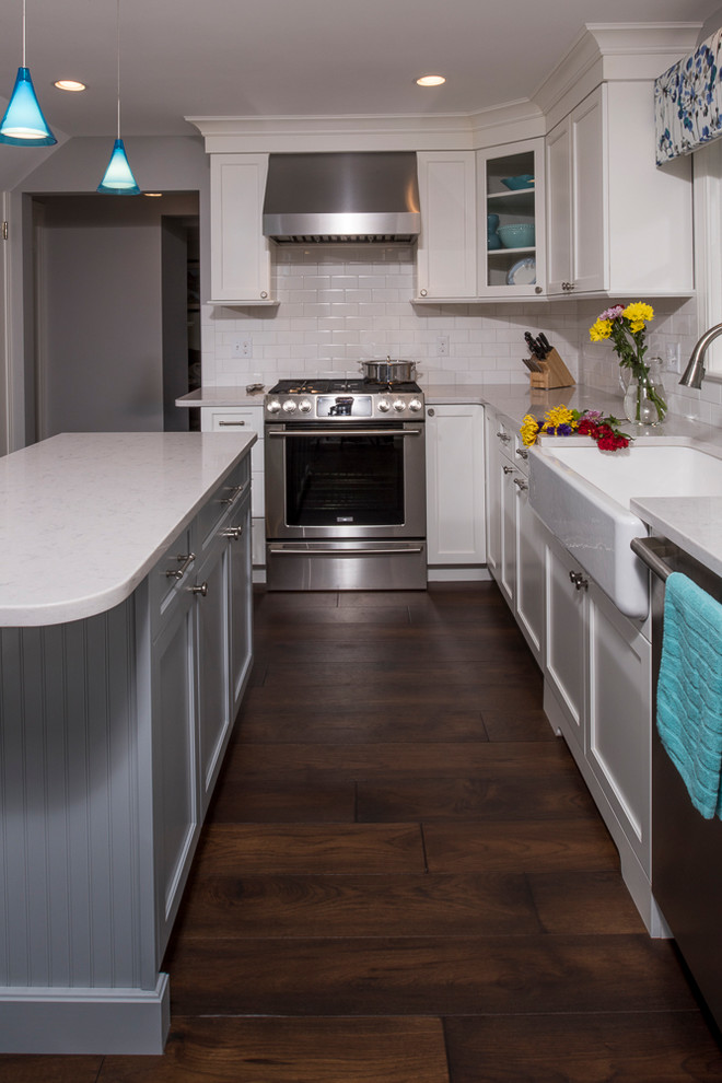 Transitional l-shaped dark wood floor and brown floor kitchen photo in Boston with a farmhouse sink, flat-panel cabinets, white cabinets, quartz countertops, white backsplash, ceramic backsplash, stainless steel appliances and an island