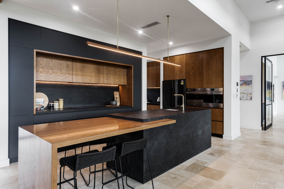 This is an example of a large industrial open plan kitchen in Adelaide with a farmhouse sink, black cabinets, quartz benchtops, black splashback, stone slab splashback, black appliances, travertine floors, with island, beige floor and black benchtop.