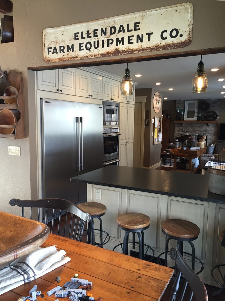 Inspiration for a mid-sized rustic l-shaped dark wood floor enclosed kitchen remodel in Minneapolis with a farmhouse sink, raised-panel cabinets, distressed cabinets, granite countertops, black backsplash, stainless steel appliances and an island