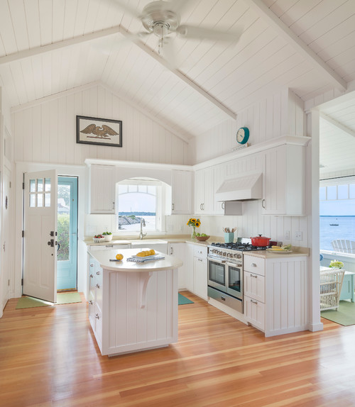 White coastal kitchen with vertical V-groove (shiplap-style) cabinet fronts and paneling, vaulted ceiling, wood floors, and a small island overlooking the water