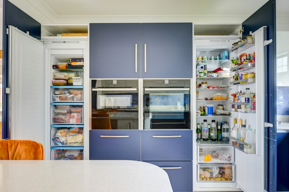 サセックスにあるお手頃価格の広いコンテンポラリースタイルのおしゃれなキッチン (一体型シンク、フラットパネル扉のキャビネット、青いキャビネット、珪岩カウンター、青いキッチンパネル、ガラス板のキッチンパネル、パネルと同色の調理設備、クッションフロア、茶色い床、白いキッチンカウンター) の写真