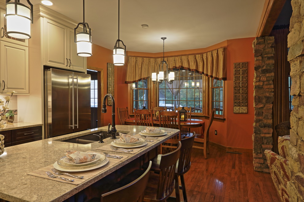 Example of a large transitional l-shaped light wood floor eat-in kitchen design with an undermount sink, raised-panel cabinets, brown cabinets, quartz countertops, beige backsplash, ceramic backsplash, stainless steel appliances and an island