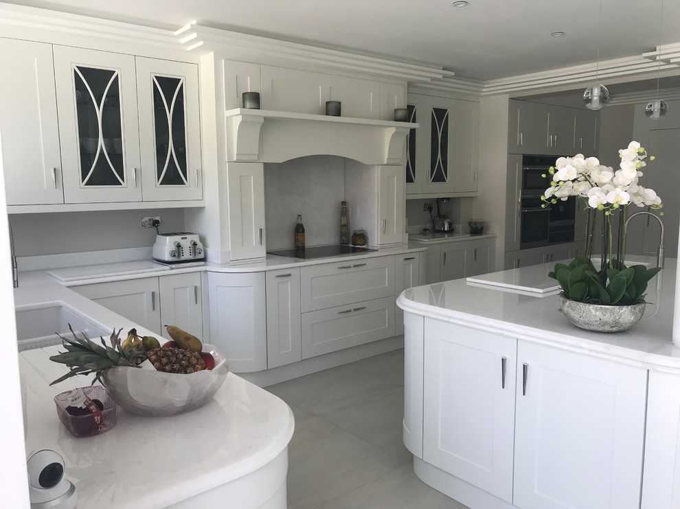 This is an example of a large contemporary open plan kitchen in Hertfordshire with a built-in sink, shaker cabinets, white cabinets, quartz worktops, white splashback, stone slab splashback, stainless steel appliances, porcelain flooring, an island, white floors and white worktops.